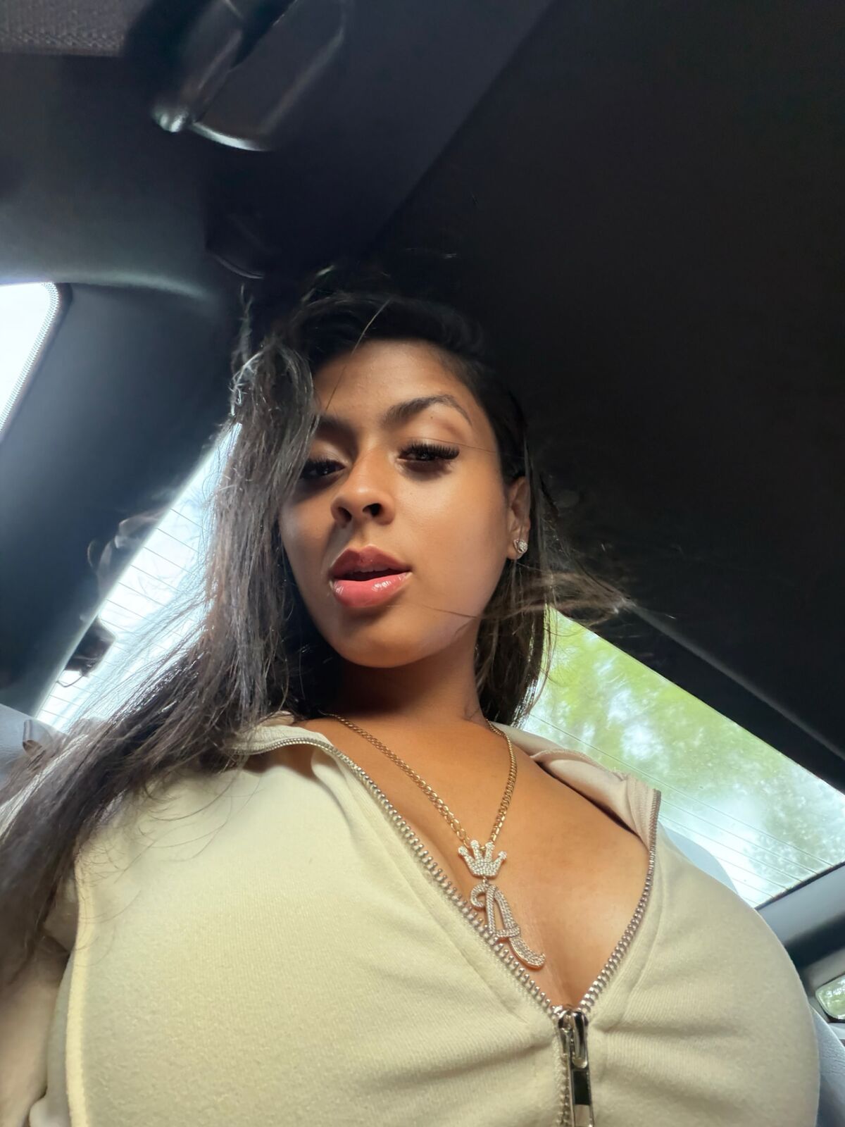 Close-up car selfie of LA Real Ash with long dark hair, beige zip-up top and necklace